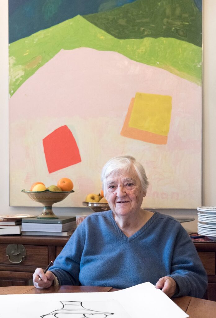 Donna anziana seduta ad un tavolo. La donna ha di fronte dei fogli illustrati e alle sue spalle una tela dipinta. sulla tela ci sono delle montagne stilizzate in verde e due quadrati colorati, uno rosso e uno giallo, su uno sfondo bianco.